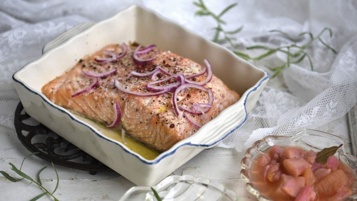 Lachs in einer eckigen weißen Auflaufform. Garniert mit roten Zwiebeln und Pfeffer. Rechts daneben befindet sich eine kleine Glasschale mit Rhabarberkompott. Frische Estragonzweige sind dekorativ um die Speisen drapiert.