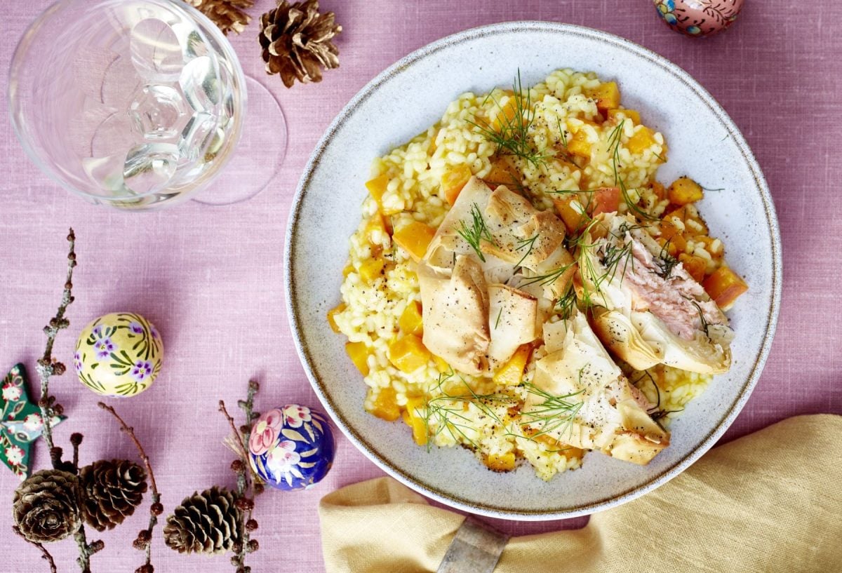Lachs-Päckchen auf Kürbis-Risotto, garniert mit frischem Dill und serviert auf einem weißen Teller auf rosanem Untergrund.