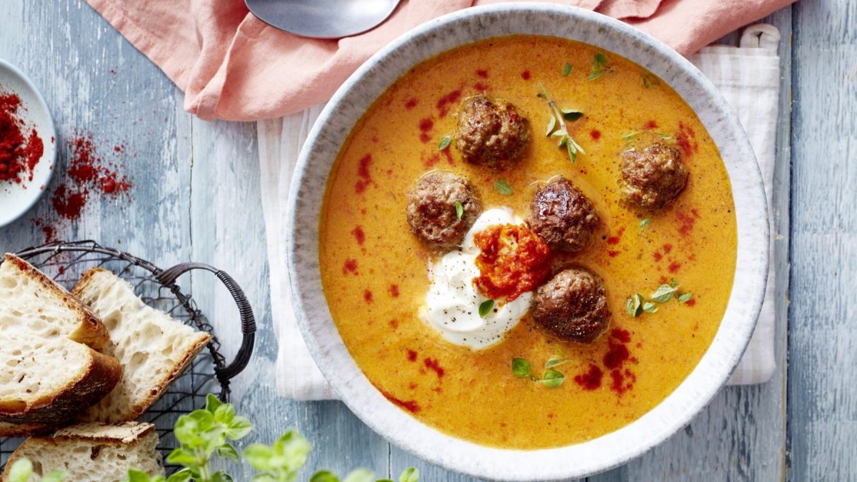 Paprikasuppe mit Hackbällchen, garniert mit Schmand, Ajvar und frischem Oregano und serviert in einer Suppenschüssel auf einem rustikalen Holztisch.