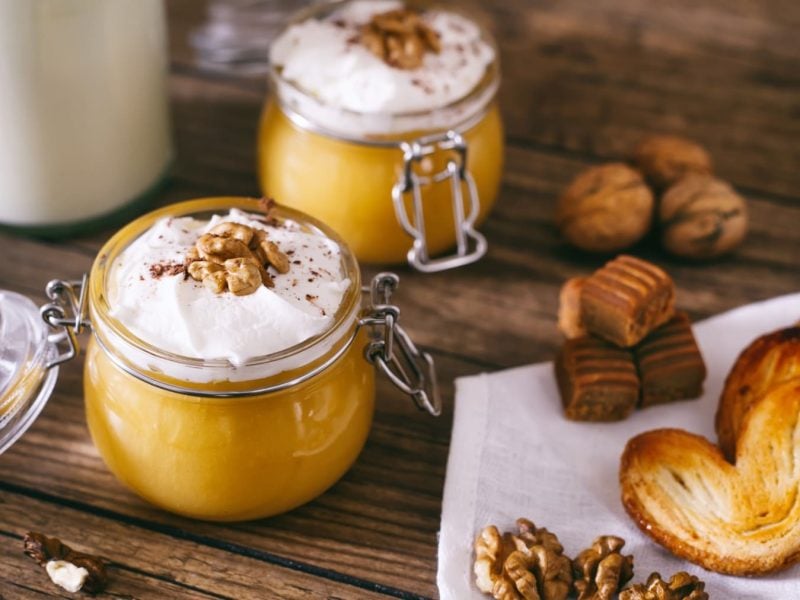 Zwei Einmachgläser mit Kürbis-Walnuss-Shake mit Sahne, daneben verschiedene Kekse vor Holzhintergrund.