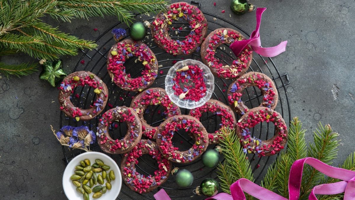 Mehrere Schokoladenkringel mit gefriergetrockneten Himbeeren auf einem Gitter neben Tannenzweigen, Pistazien, Weihnachtskugeln und pinkem Band vor grauem Hintergrund