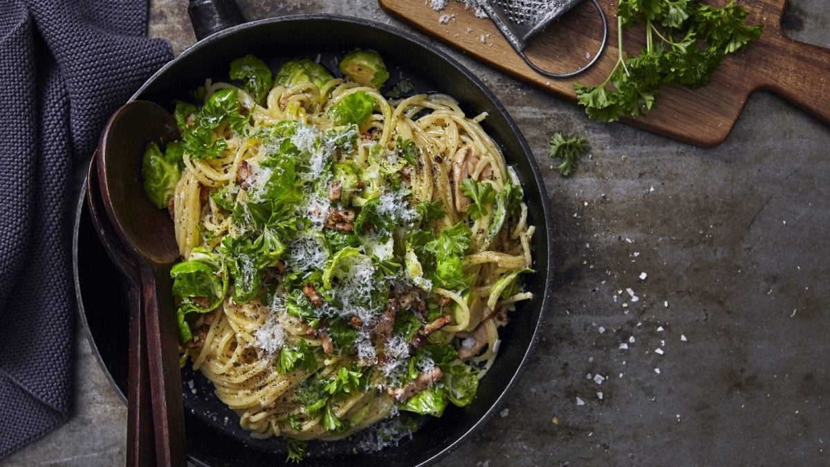 In einer Pfanne sind Spaghetti mit Rosenkohl, Speck, Parmesan und Petersilie angerichtet. Sie sind mit Käse bestreut.