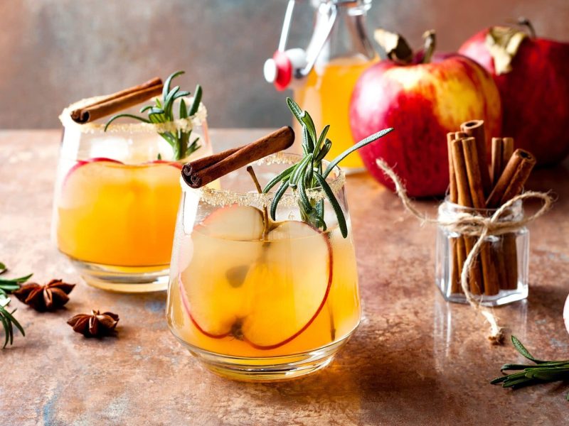Zwei Gläser Apple Cider Margarita mit Rosmarin, Zimtstangen und Äpfeln.