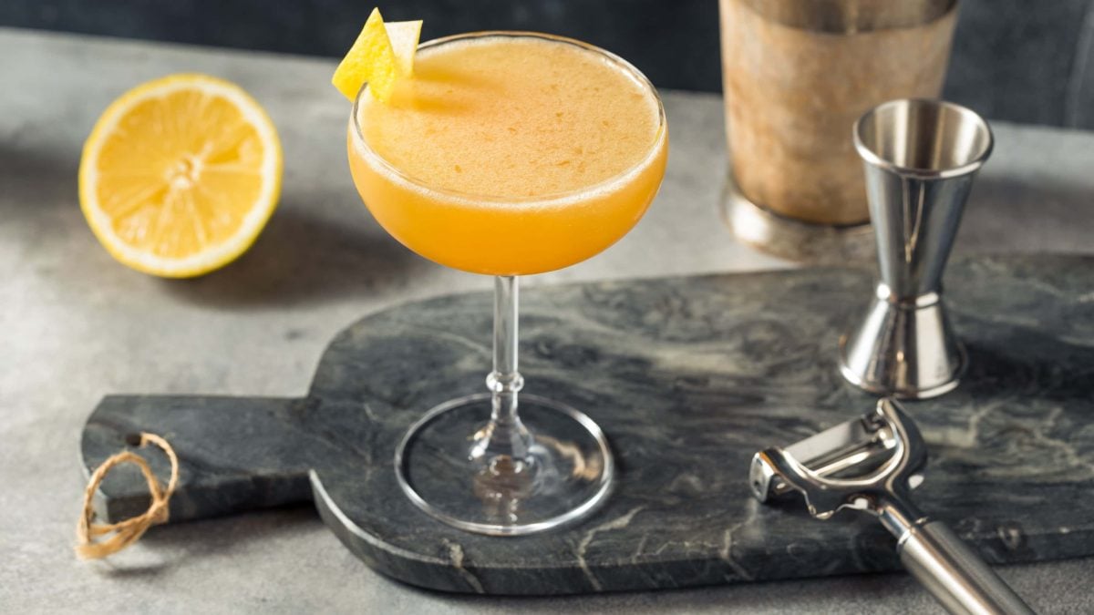 Apple Jack Rabbit Calvados Cocktail in Cocktailschale mit Zitronenzeste auf Marmorplatte mit Barmaß und Zestenreißer. Im Hintergrund halbe Orange und Shaker. Frontalansicht.