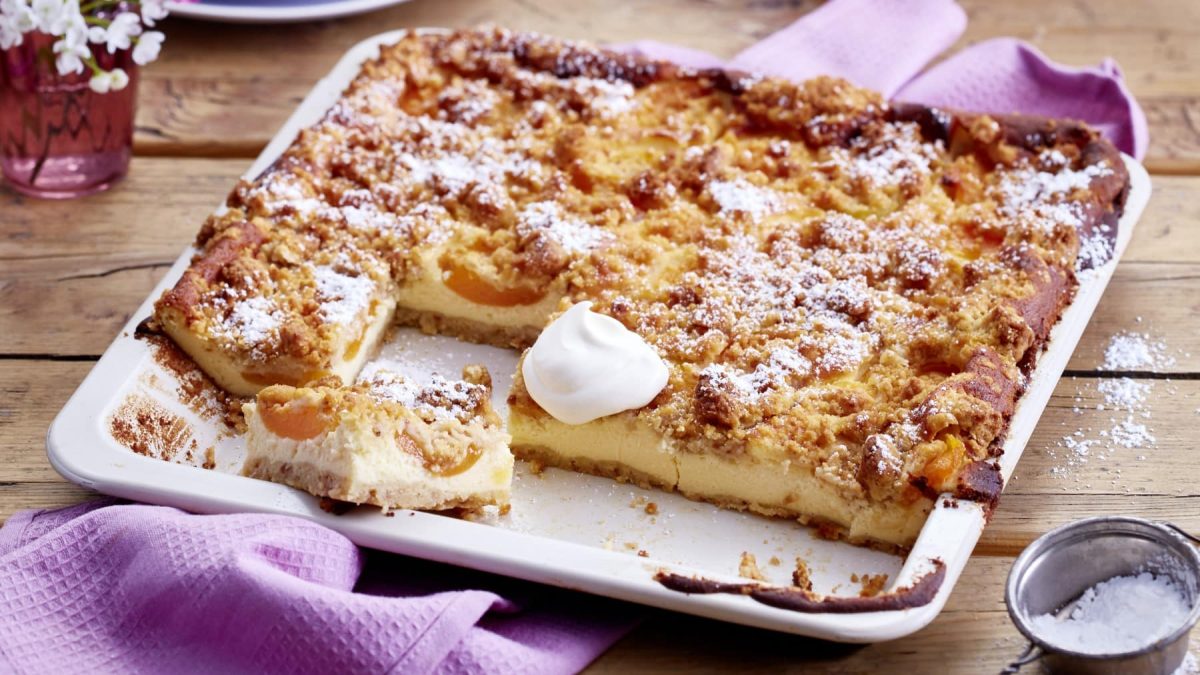 Aprikosen-Schmand-Kuchen im Blech auf einem lila Tuch vor Holzhintergrund.