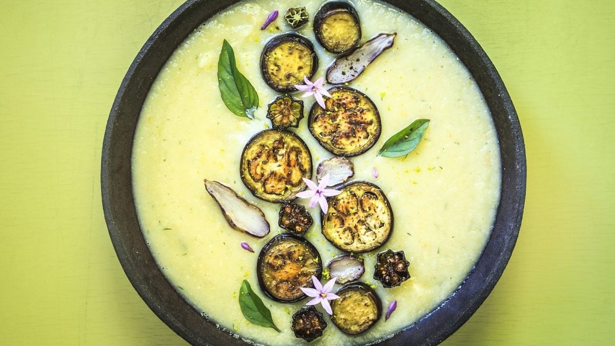 Auberginensuppe mit getrocknetem Gemüse, Thai-Basilikum und gerösteten Auberginenscheiben, in einer dunklen Schale auf gelbem Untergrund serviert.