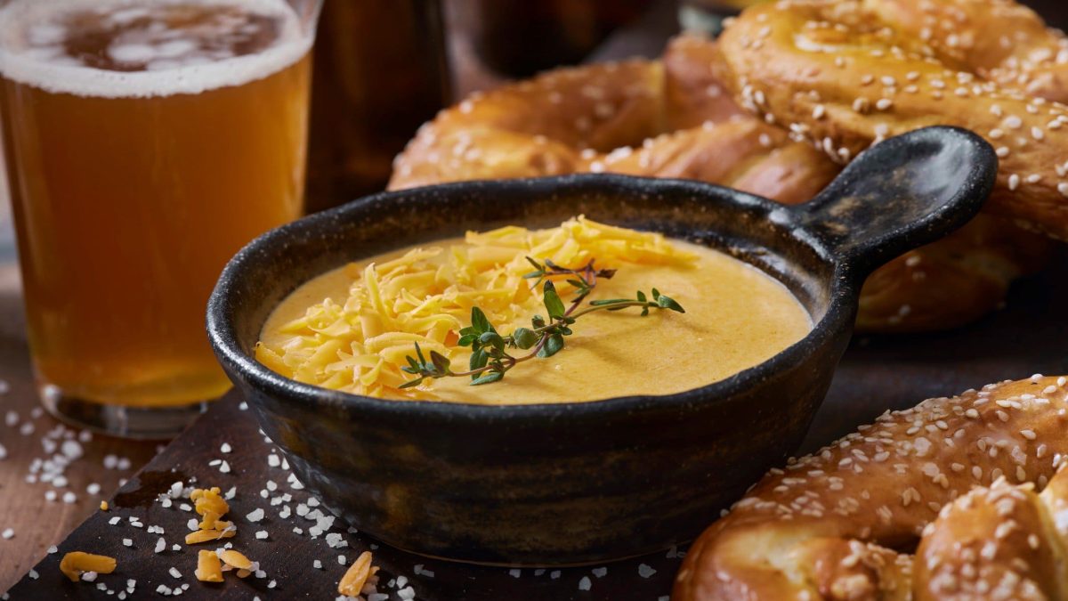 Beer Cheese Dip in einer schwarzen Schale auf Granitplatte. Im Bild außerdem Bretzeln und ein Glas Bier. Grobes Salz und Thymian im Bild verteilt. Frontalaufnahme..