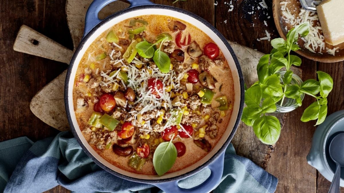 Draufsicht: Blauer gusseiserner Topf mit Bergsteiger-Pizza-Eintopf mit Tomaten, Pilzen, Mais und grüner Paprika. Garniert mit Käse und Basilikum. Angerichtet auf einer rustikalen Holzoberfläche, daneben geriebener Parmesan und frischer Basilikum.