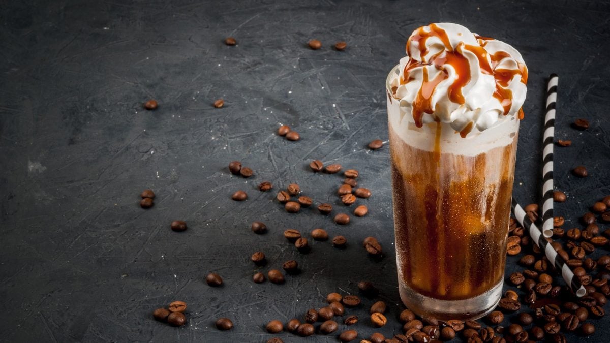 EIn Glas Caramel Macchiato mit Schlagsahne und Karamellsauce, daneben Kaffeebohnen und Strohhalme.