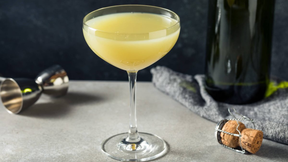 Death in the Afternoon Cocktail mit Absinth und Champagner von Ernest Hemingway in Cocktailschale auf grauem Tisch. Im Hintergrund eine Flasche Champagner und ein Barmaß. Vorne Korken. Frontalansicht.