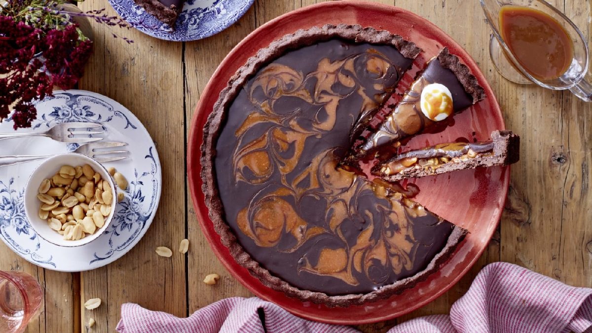 Draufsicht: Angeschnittene Torte mit Karamell-Schokoladen-Marmorierung. Serviert auf einem roten Tortenteller. Dekoriert mit extra Erdnüssen und einem Saucenspender, der flüssiges Karamell enthält.