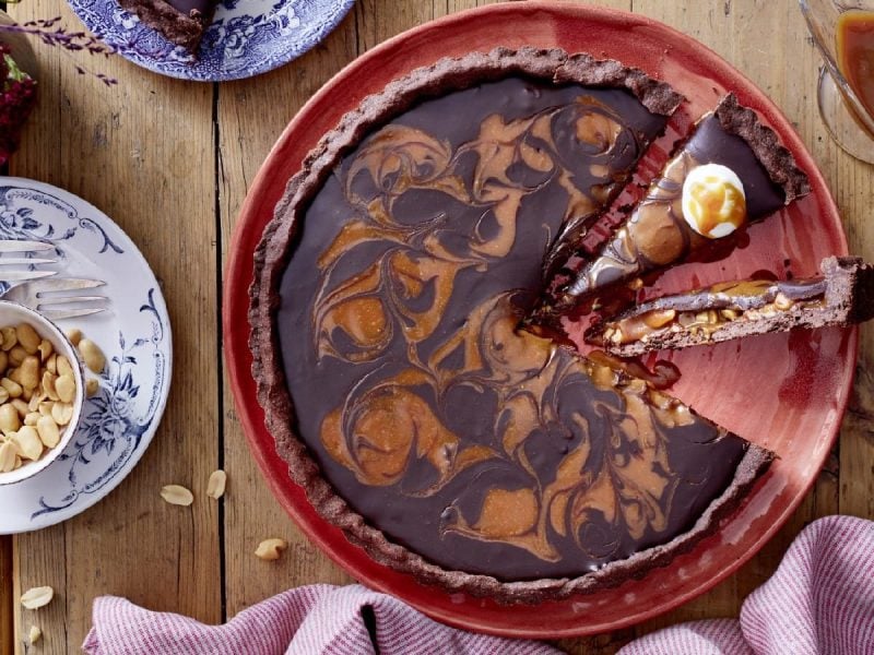 Draufsicht: Angeschnittene Torte mit Karamell-Schokoladen-Marmorierung. Serviert auf einem roten Tortenteller. Dekoriert mit extra Erdnüssen und einem Saucenspender, der flüssiges Karamell enthält.
