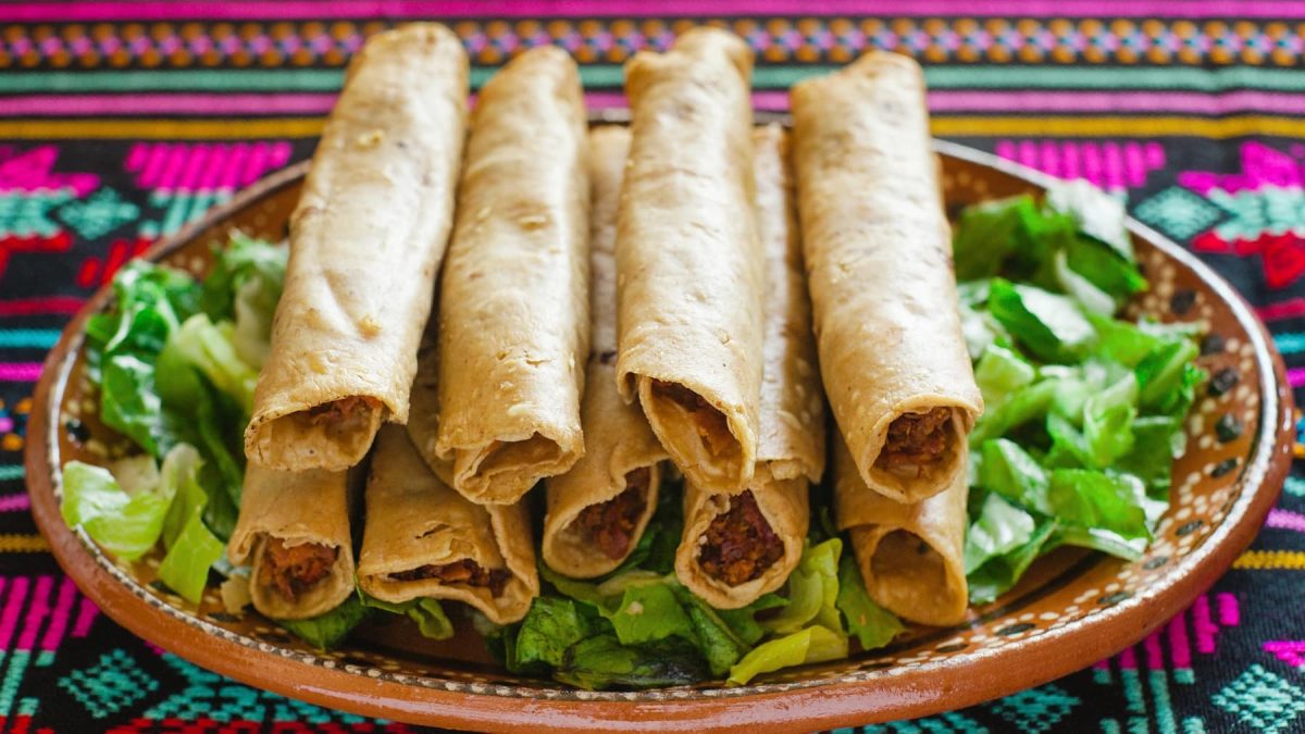 Mehrere gerollte Hühnertacos auf einem braunen Teller mit salat darunter, auf einer bunten Decke.