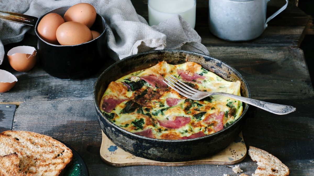 Eine Frittata mit Spinat und Schinken in einer gusseisernen Pfanne, daneben Eier und geröstetes Brot.