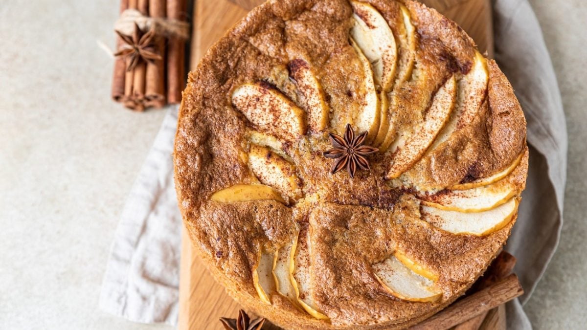 Draufsicht: ein runder Apfelkuchen, auf dem ein Sternanis liegt. Der Gewürz-Apfelkuchen liegt auf einem Bretten, daneben liegen mehrere Stangen Zimt und ein weiteres Sternanis.