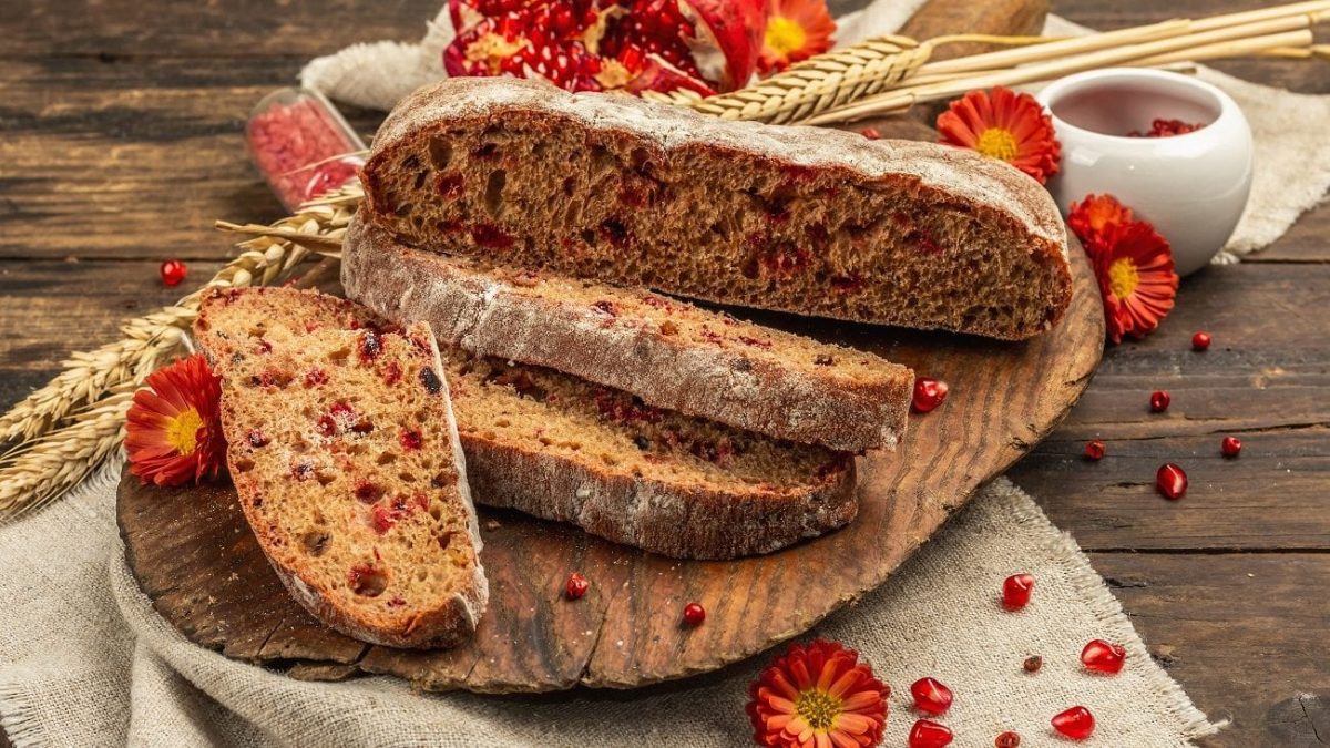 Frontalsicht: Ein Granatapfelbrot liegt auf einem Leinentuch, welches auf einem Untergrund aus Holz liegt. Drumherum liegen ein aufgebrochener Granatapfel sowie getrocknete Blüten.
