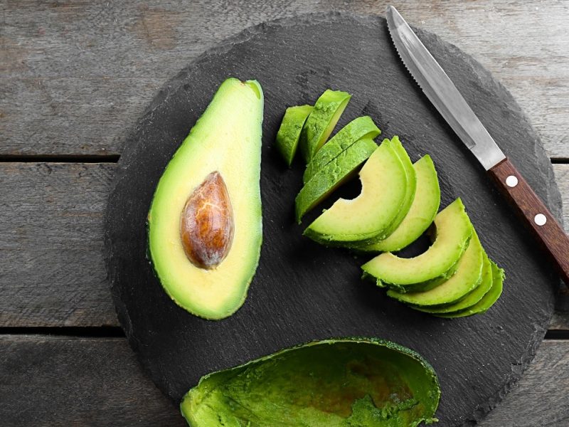 Draufblick: eine Avocadohälfte mit einem kern ligt auf einen Schieferbrett und dieses auf einem dunklen Untergund. Daneben liegt Avocadoscheiben.
