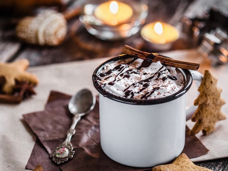 Frontalsicht: EIn weihnachtliche gedeckter Tisch mit Kerzen, Kekesen und eine Blechtasse, in der ein hesses Getränk mit einer Sahnehaube steht. Es handelt sich um eine heisse Lebkuchen-Schokolade.
