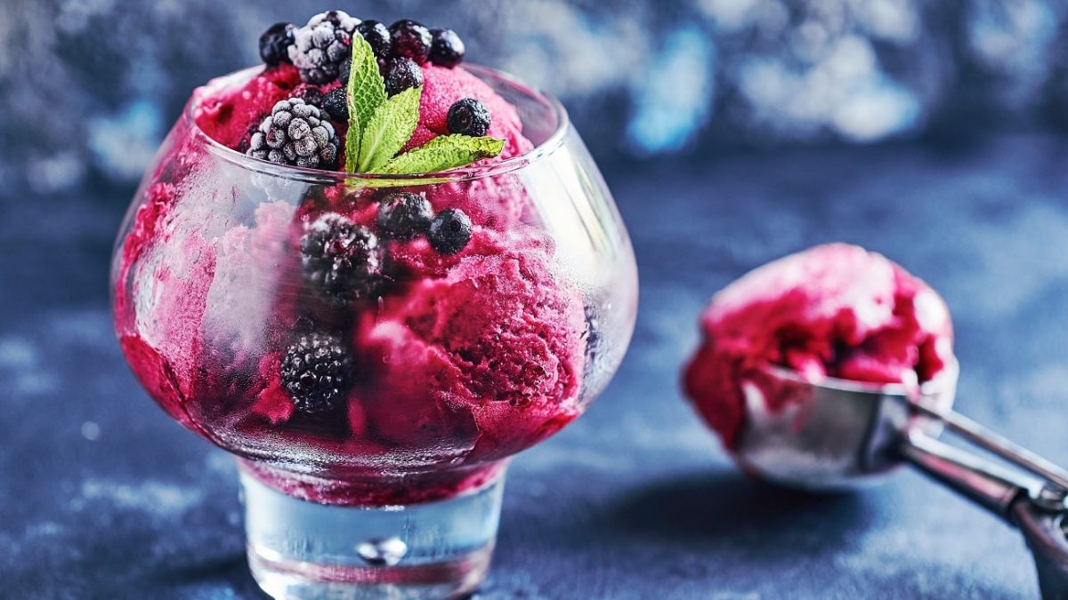 Frontalsicht: Ein Glas mit einem Johannisbeer-Brombeer-Sorbet mit Prosecco. Daneben liegt ein Eisportionierer. der Untergrund ist dunkel.
