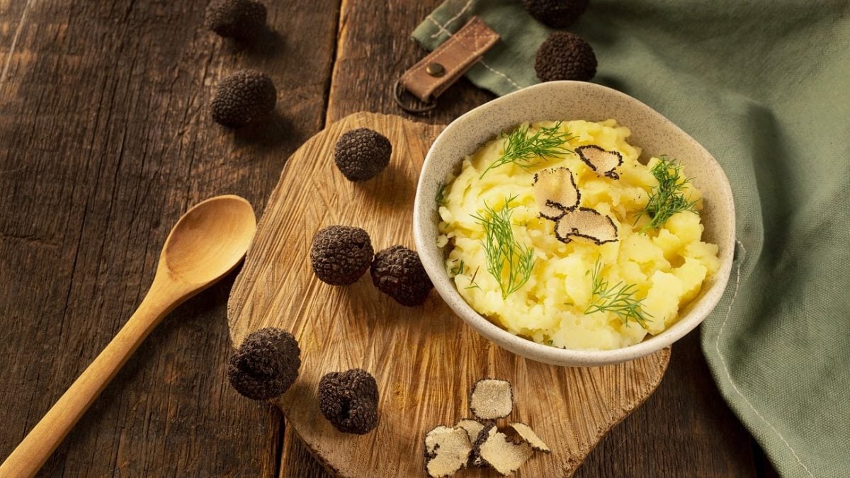 Draufsicht: Ein Topf mit Kartoffelpüree mit gehobeltem Trüffel steht auf einem Holzbrettchen. Daneben liegen ein Kochlöffel aus Holz und dunkle, runde Trüffel.