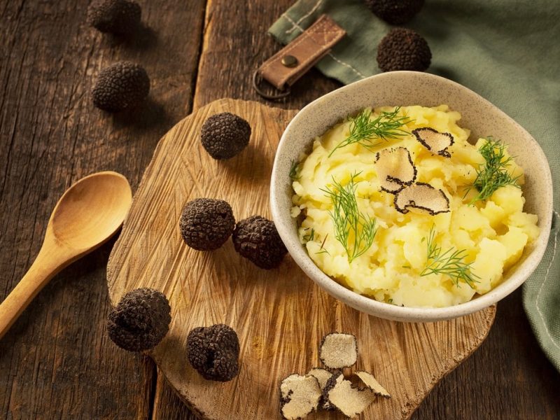 Draufsicht: Ein Topf mit Kartoffelpüree mit gehobeltem Trüffel steht auf einem Holzbrettchen. Daneben liegen ein Kochlöffel aus Holz und dunkle, runde Trüffel.