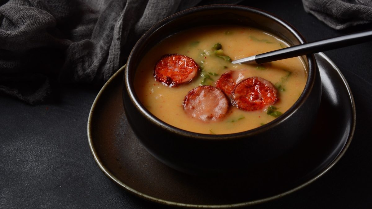 Eine schwarze Schüssel auf dunklem Hintergrund. Darin die Kartoffelsuppe mit ein paar Scheiben gebratener Chorizo.