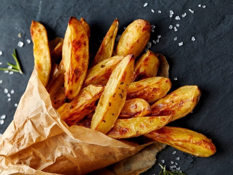 Eine Papiertüte Kohlrabi-Fritten mit Salz und Rosmarin auf dunklem Untergrund.