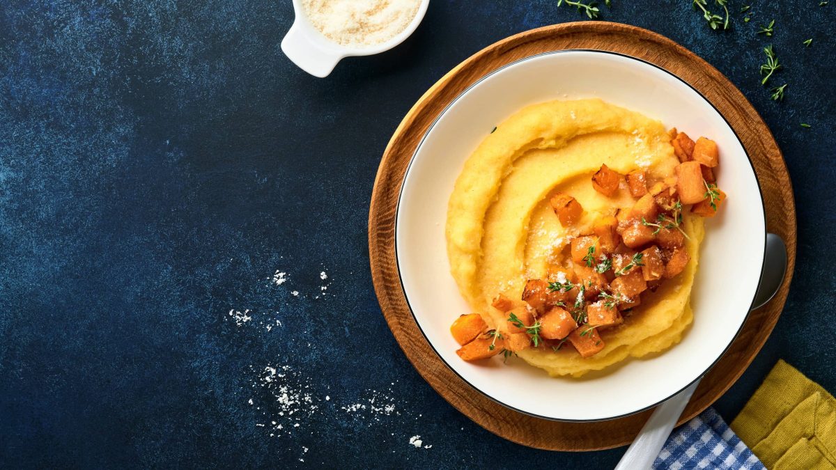 Ein weißer Teller Kürbis-Polenta auf einem dunkelblauem Untergrund. Darüber eine Schale Parmesan und etwas Thymian.