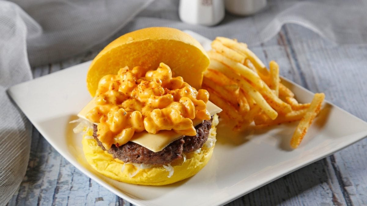 Mac'n Cheese Burger mit Coleslaw auf weißem Teller mit Pommes auf hellem Untergrund. Frontalansicht.