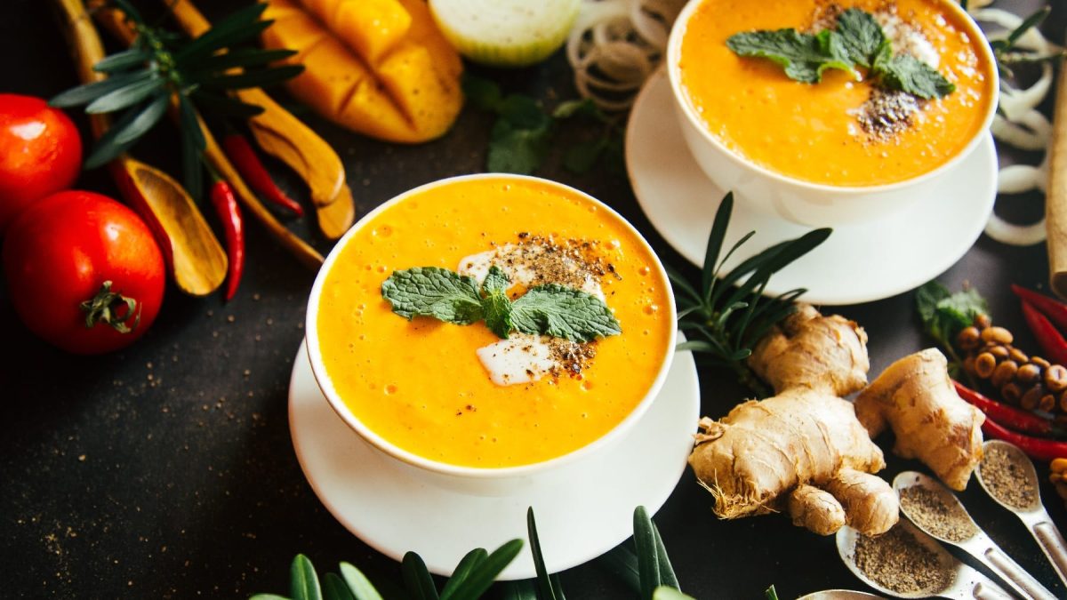 Zwei Suppenschalen mit der gelben Suppe auf dunklem Untergrund und allerlei Zutaten und Gewürzen daneben.