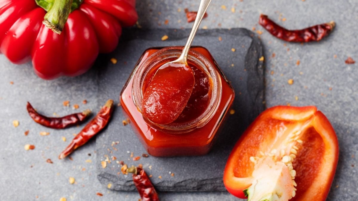 Draufsicht: In einem Glas ist eine Rote, cremige Masse. Auf dem Glas liegt ein teelöffel mit der Masse. Drum herum liegt eine ganze Paprika, eine halbe Paprika sowie Chilischoten.