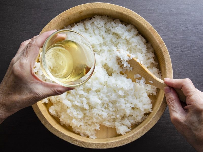 Perfekter Sushi-Reis, zubereitet mit Reisessig und Zucker, vermengt in einer Holzschale auf dunklem Untergrund.