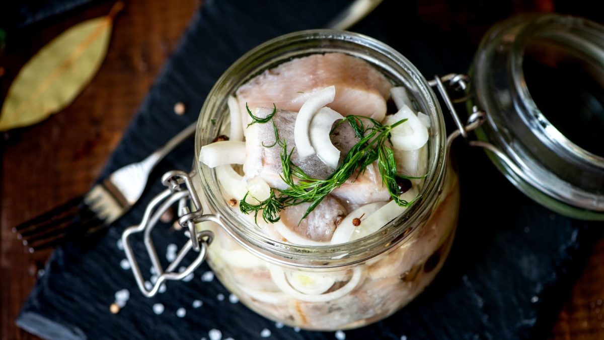 Heringstücke mit Zwiebelscheiben und Pfefferkörnern in einem Bügelglas auf schwarzem Untergrund.