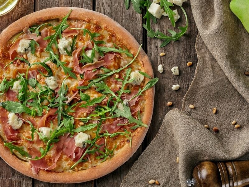 Draufsicht: Eine Pizza mit Schinken, Birne und Gorgonzola liegt auf einem Untergund aus Holz. Daneben liegen Birnen, Käsewürfel, Pinienkerne, eine Pfeffermühle und ein Tuch aus Leinen.