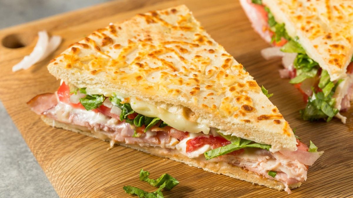 Ein Stück Pizza-Sandwich mit Schinken, Käse und Tomaten auf einem hellen Holzschneidebrett.