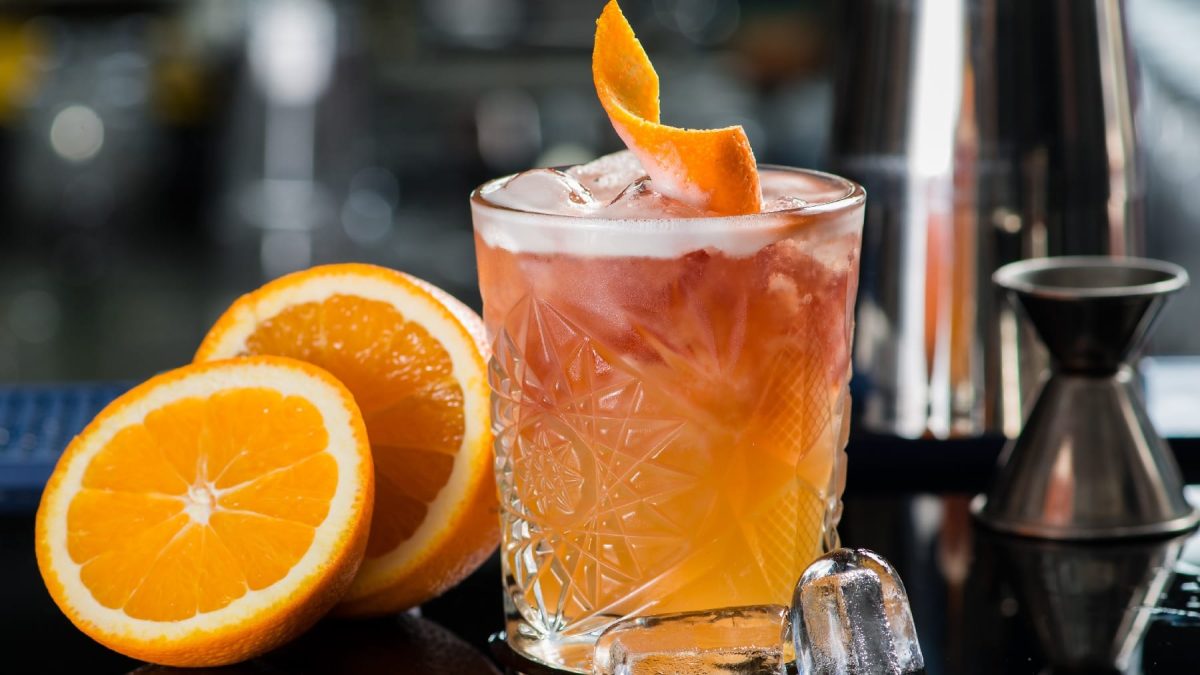 Der Cocktail Prince of Wales an einer Bartheke mit einer halbierten Orange und einer ganzen Menge Barequipment im Hintegrund.