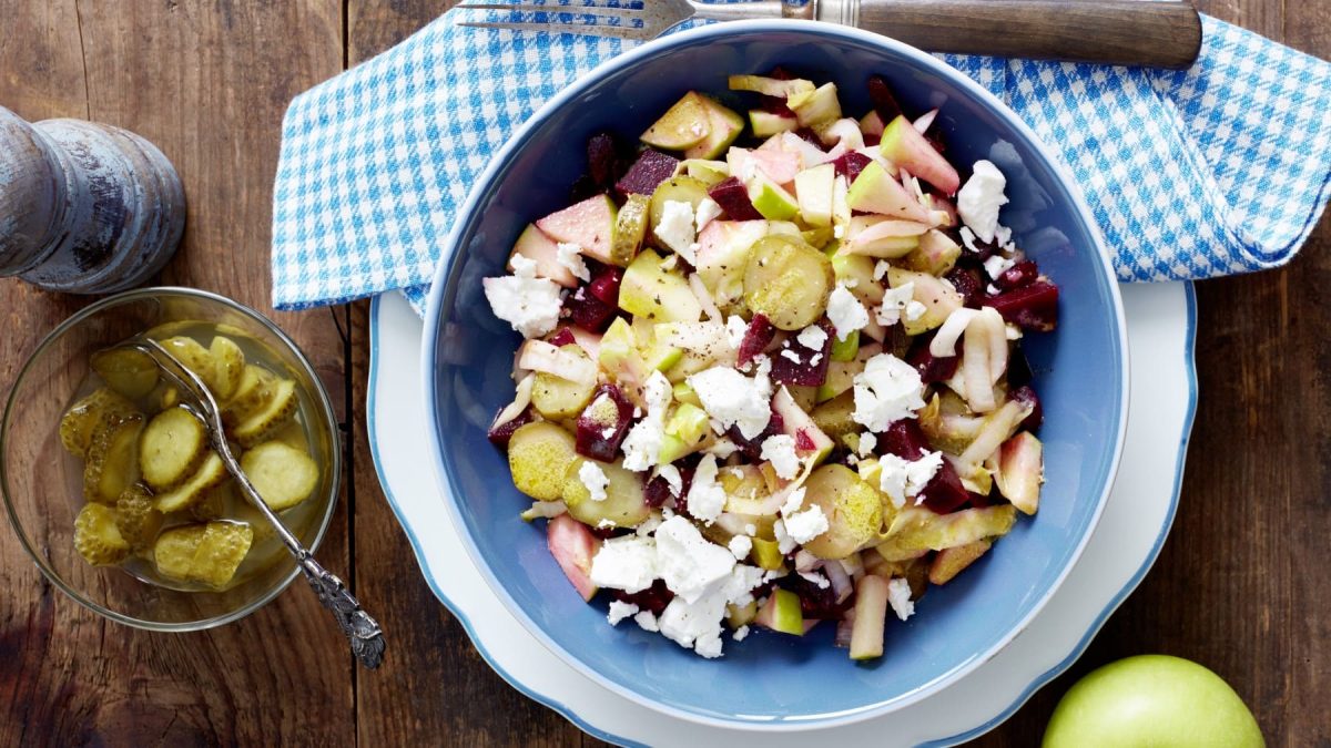 Rote-Bete-Salat mit Feta in blauer Schüssel, daneben Gewürzmühle, saure Gurken und ein grüner Apfel vor Holzhintergrund.