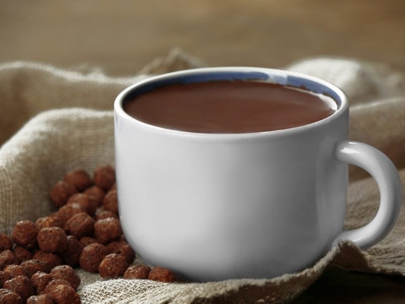 Frotansicht: eine helle Tasse mit einer dunklen Flüssigkeit steht auf sachleinen. Neben an lieben ein paar Schokodrops.