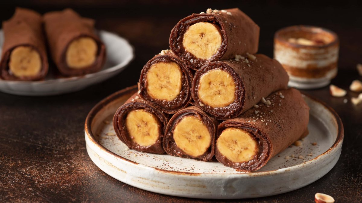 Mehrere gerollte Schoko-Crepes gefüllt mit Banane auf einem grauen Teller und braunem Untergrund.