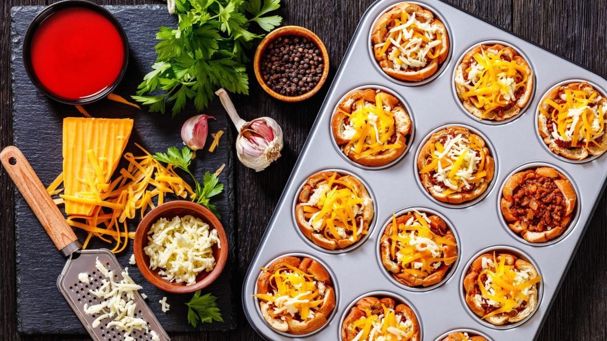 Sloppy Joes Muffins in Muffinform mit Käse. Daneben Zutaten und Käsereibe. Draufsicht.