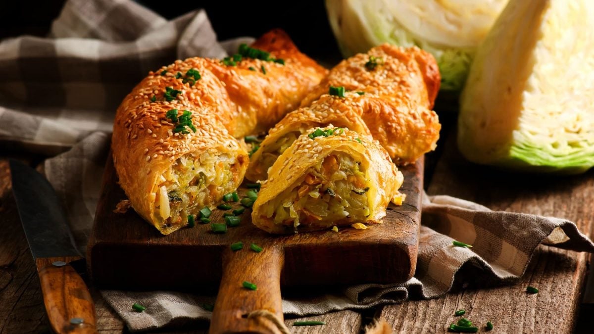 Ein Schneidebrett mit dem Strudel gefüllt mit Kohl, Kartoffeln und Karotten auf einem rustikalen Holztisch.