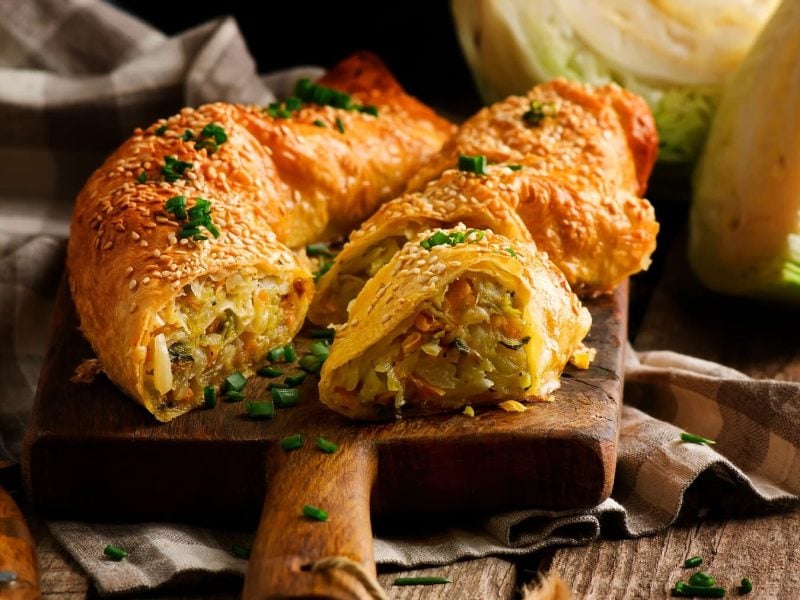 Ein Schneidebrett mit dem Strudel gefüllt mit Kohl, Kartoffeln und Karotten auf einem rustikalen Holztisch.