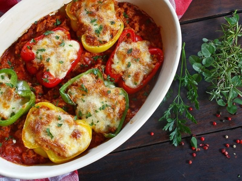 Draufsicht: Eine ofenfeste Form mit gefüllten Paprika-Hälften, die in einer Tomatensauce liegen. daneben liegen Kräuter.