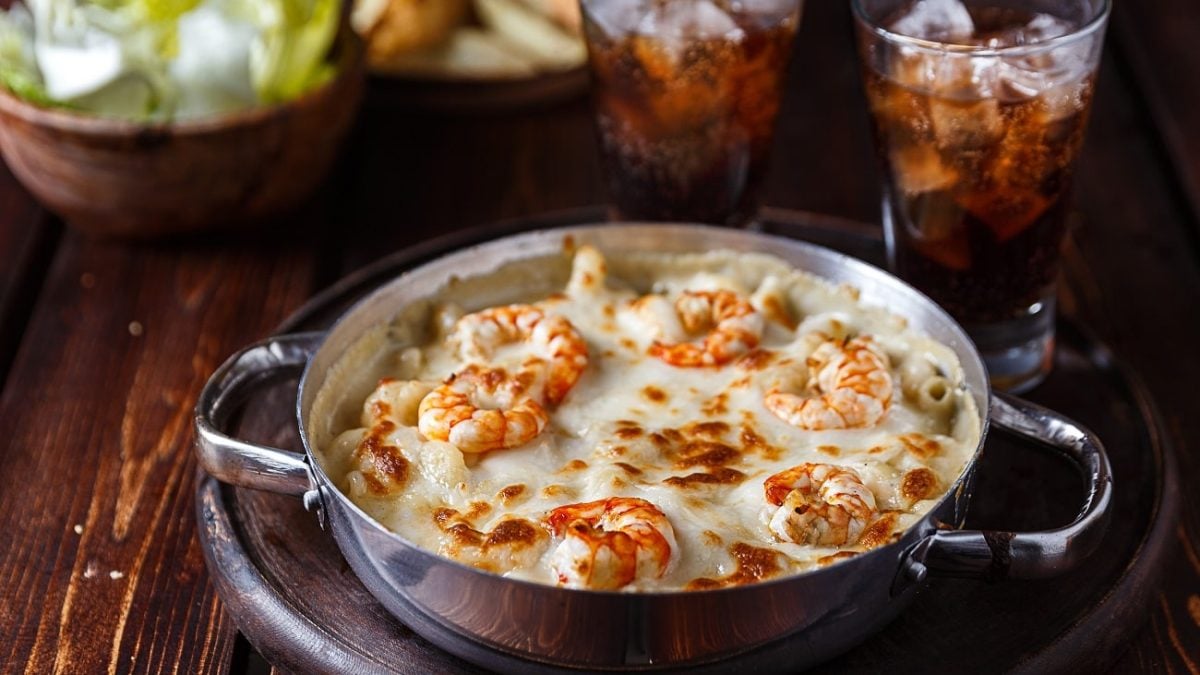 Schräge Ansicht von oben: eine dunkle Pfanne mit Überbackener Pasta mit Shrimps steht auf einem dunklen Untergrund aus Holz. Dahinter stehen zwei Gläser mit einer dunklen Flüssigkeit und Eiswürfeln.