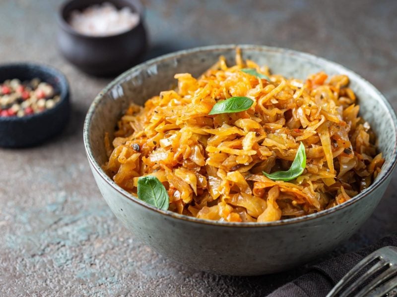 Eine Schüssel veganer Schmorkohl mit Basilikum, daneben Gabeln und Schälchen mit Gewürzen.