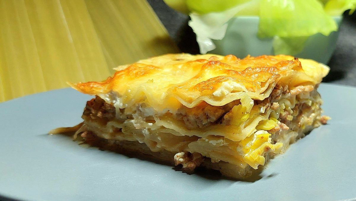 Ein grauer Teller mit einem Stück der Lasagne vor einer Schüssel mit blanchiertem Kohl und rohen Lasagneplatten.