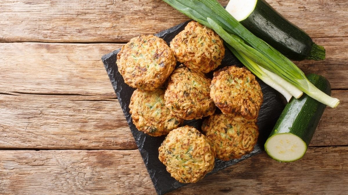 Eine dunkle Platte mit Kolokithokeftedes neben frischer Zucchini darauf.