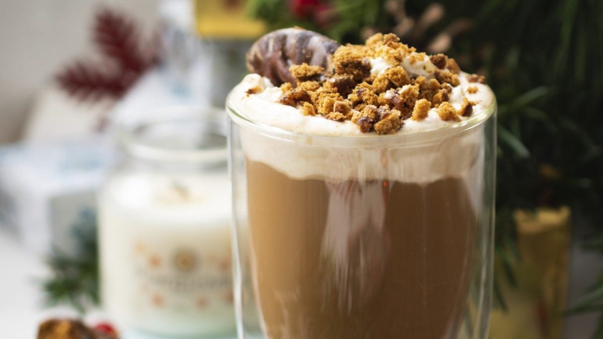 Lebkuchen-Latte mit Sahnehaube und Keks-Dekor, serviert in einem durchsichtigen Glas.