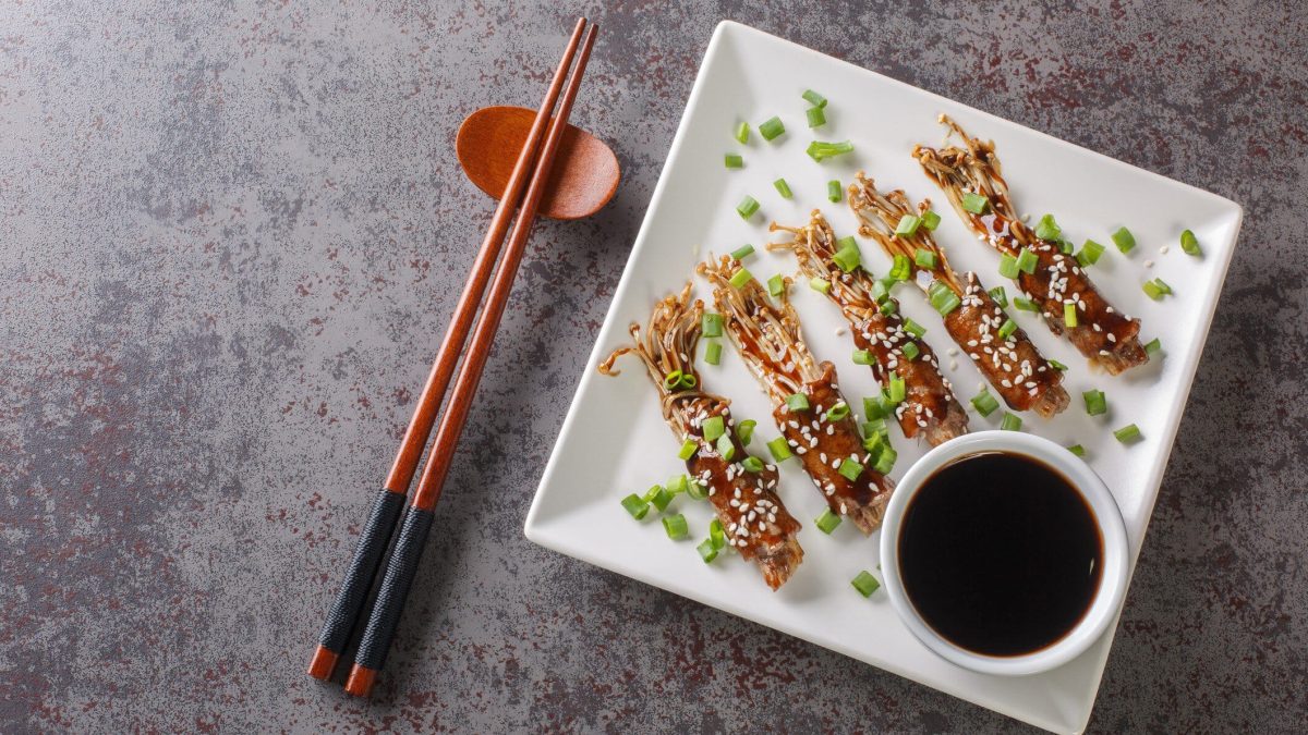 Ein weißer quadratischer Teller mit eine rkleinen Schüssel mit dunkler Sauce und den beef-Enoki-Rolls auf einem grauen Tisch.