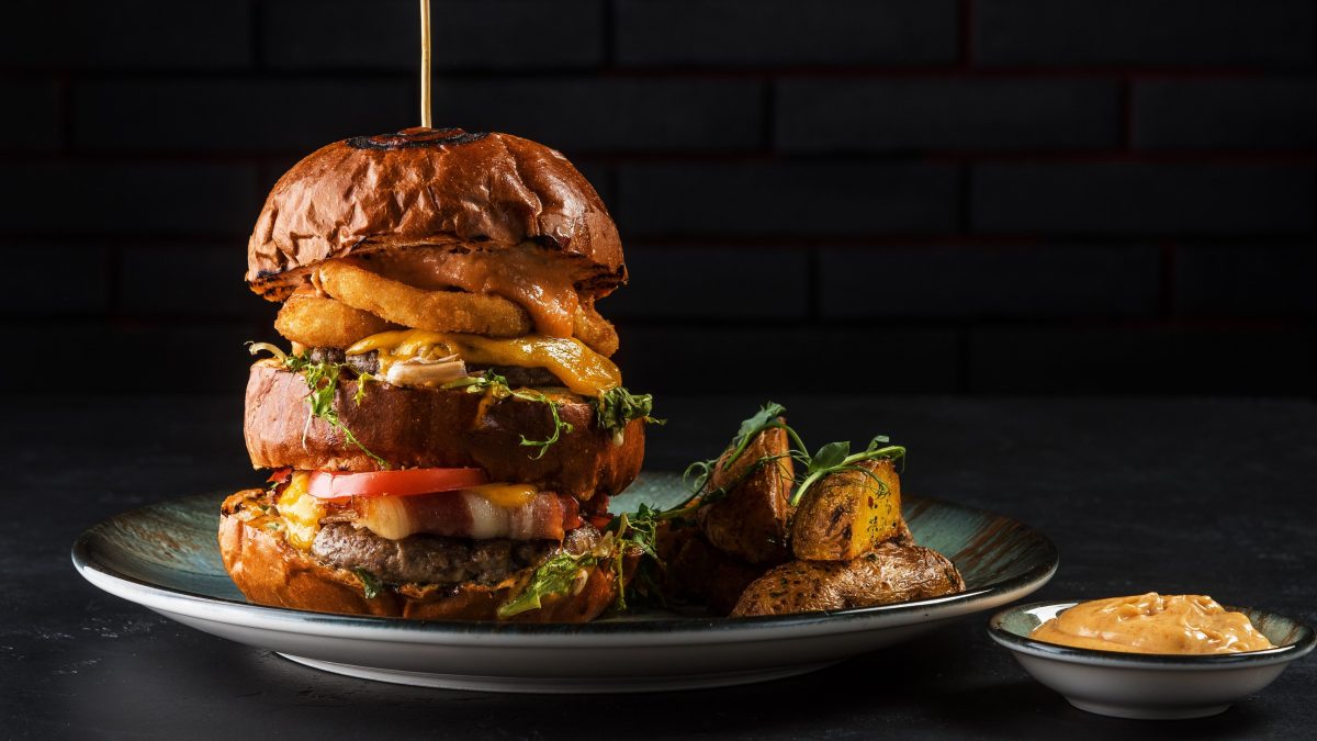 Big Boss Burger mit Wedges auf Teller und schwarzem Untergrund. Daneben Schale mit Dip. Frontalansicht.